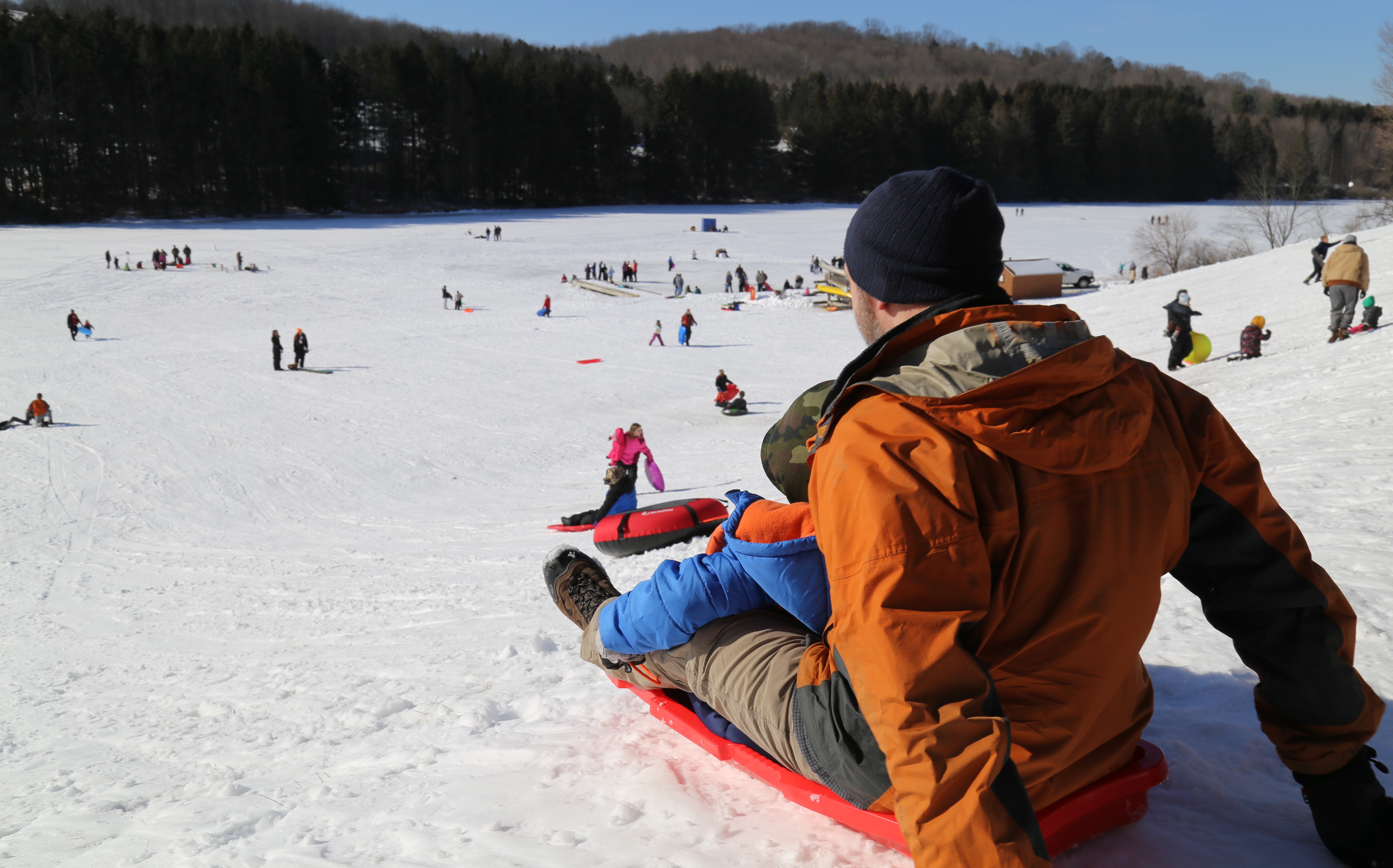 A man sledding down a steep hill with many other sledders enjoying the snowy day