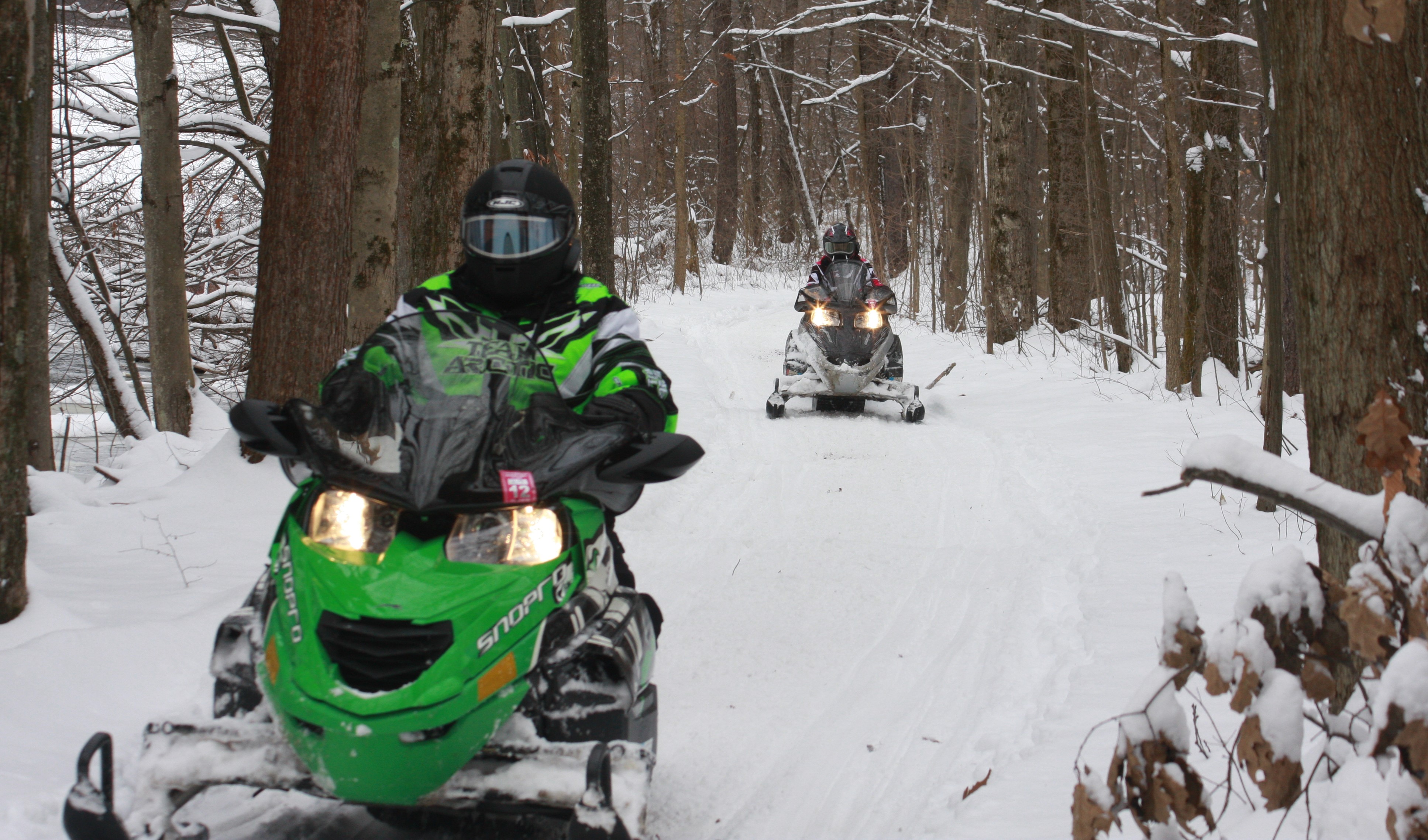 Two snowmobiles driving on a wide snowy path through the forest