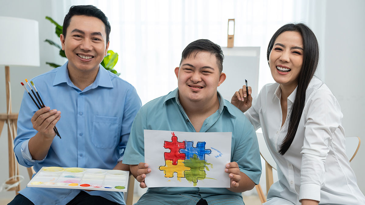 A man with autism holding a painting he painted. Two other adults are beside him holding paints and brushes.