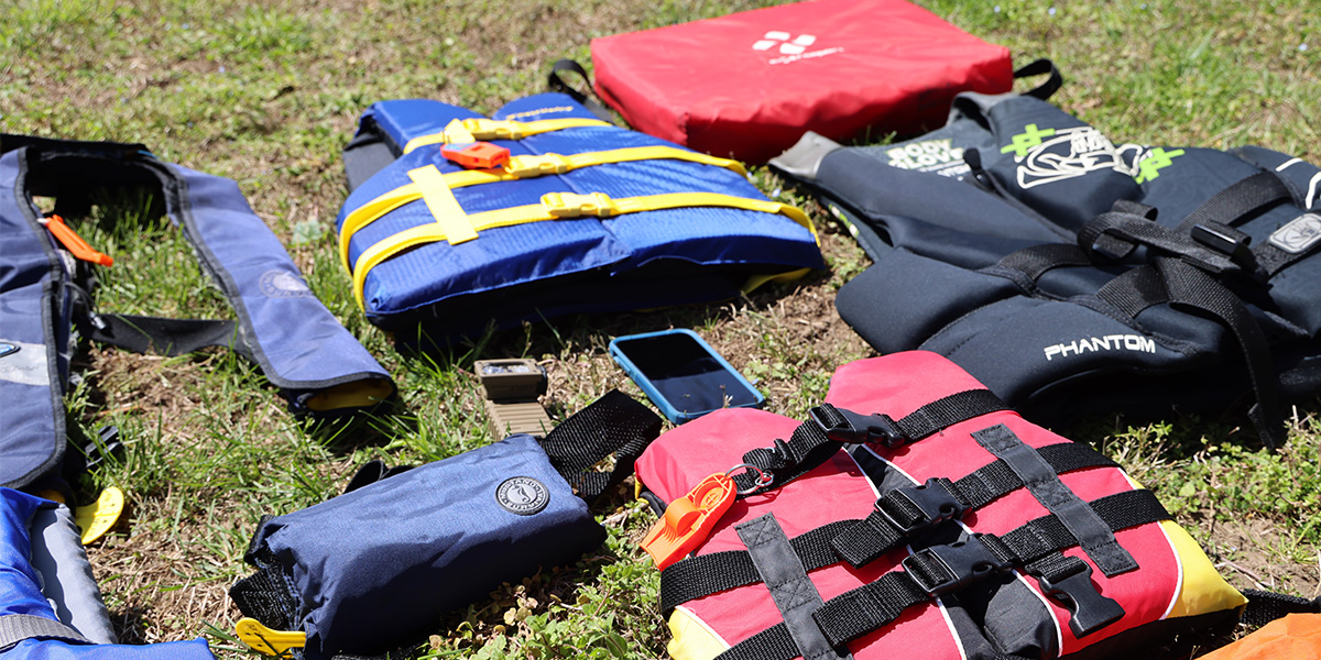 Various types of life jackets laid out on grass:  Throwable, powerboating life jacket, standard life jacket, youth life jacket and two inflatable life jackets