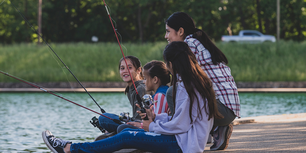 Woman with three young girls smiling and laughing while sitting on a fishing pier of a lake with their fishing rods