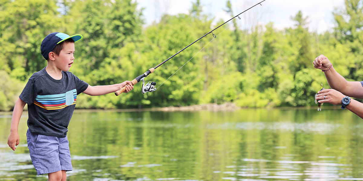 Wide angle photo of a young boy with a surprised look on his face as he holds up his fishing rod to a pair of adult's hands helping to take the fish off the hook