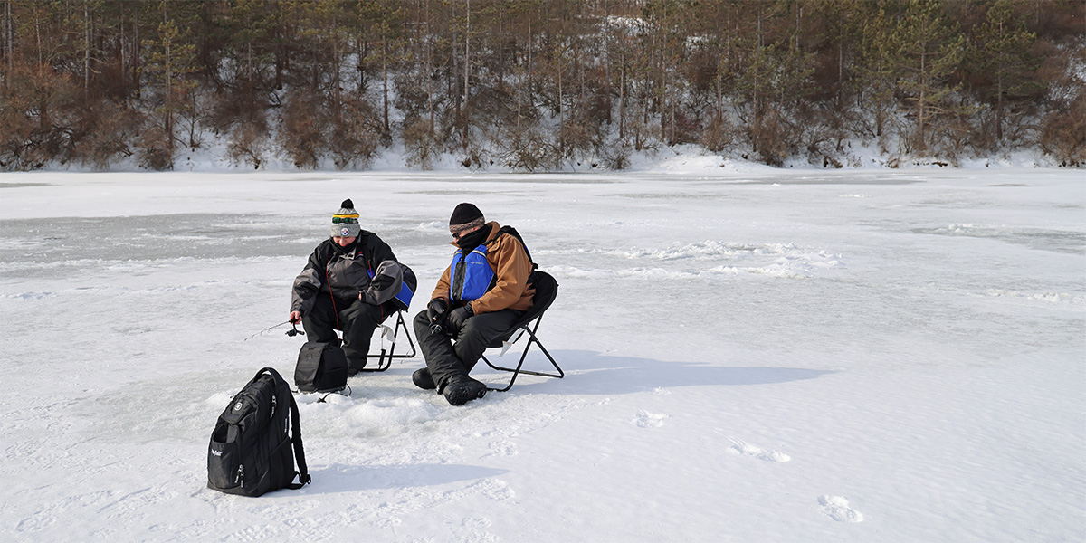 Two people bundled up in winter clothes sitting on chairs and using an ice fishing rod to fish from a small hole drilled into the frozen Nessmuk Lake.