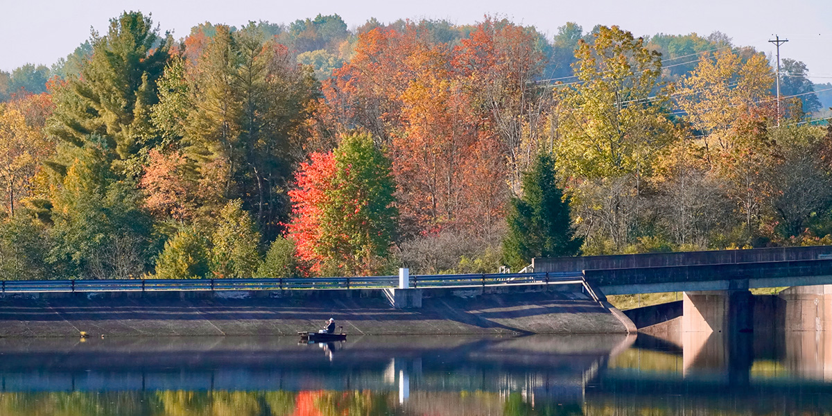 WIde shot of a boater fishing at Lyman Run State Park with the dam and fall foliage in the background.