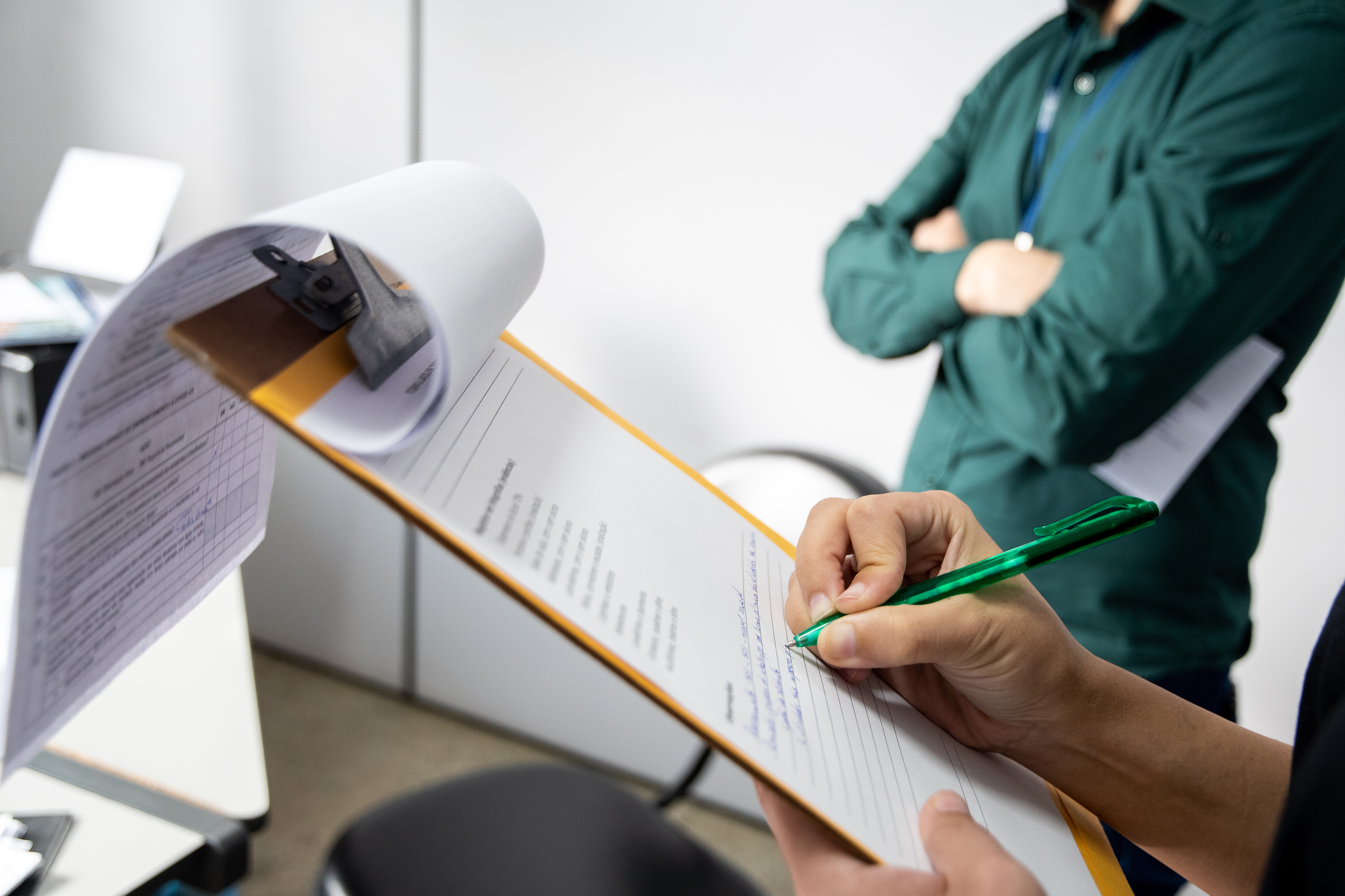 Inspector writing on a clipboard. Man in the background with arms crossed.