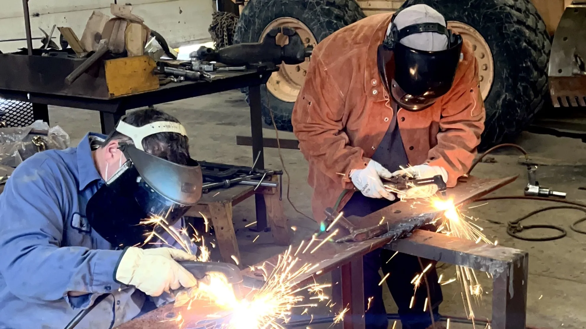 An image of PennDOT maintenance workers wearing welding masks and heavy work gloves using acetylene torches to cut notches into a grader blade.