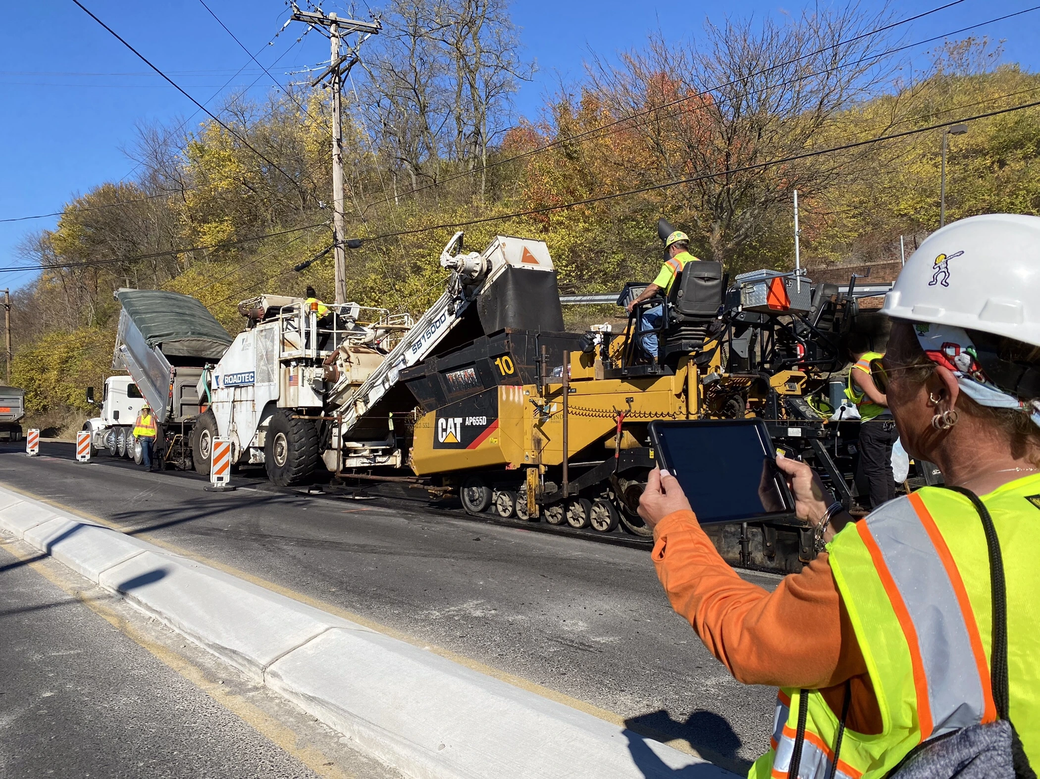 A worker holds up an electronic tablet in a work zone with construction equipment in the background.