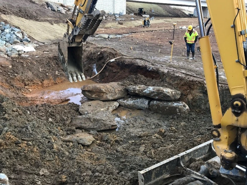 An image of a construction worker monitoring progress while a backhoe digs up and moves dirt to prepare for a roadway expansion.