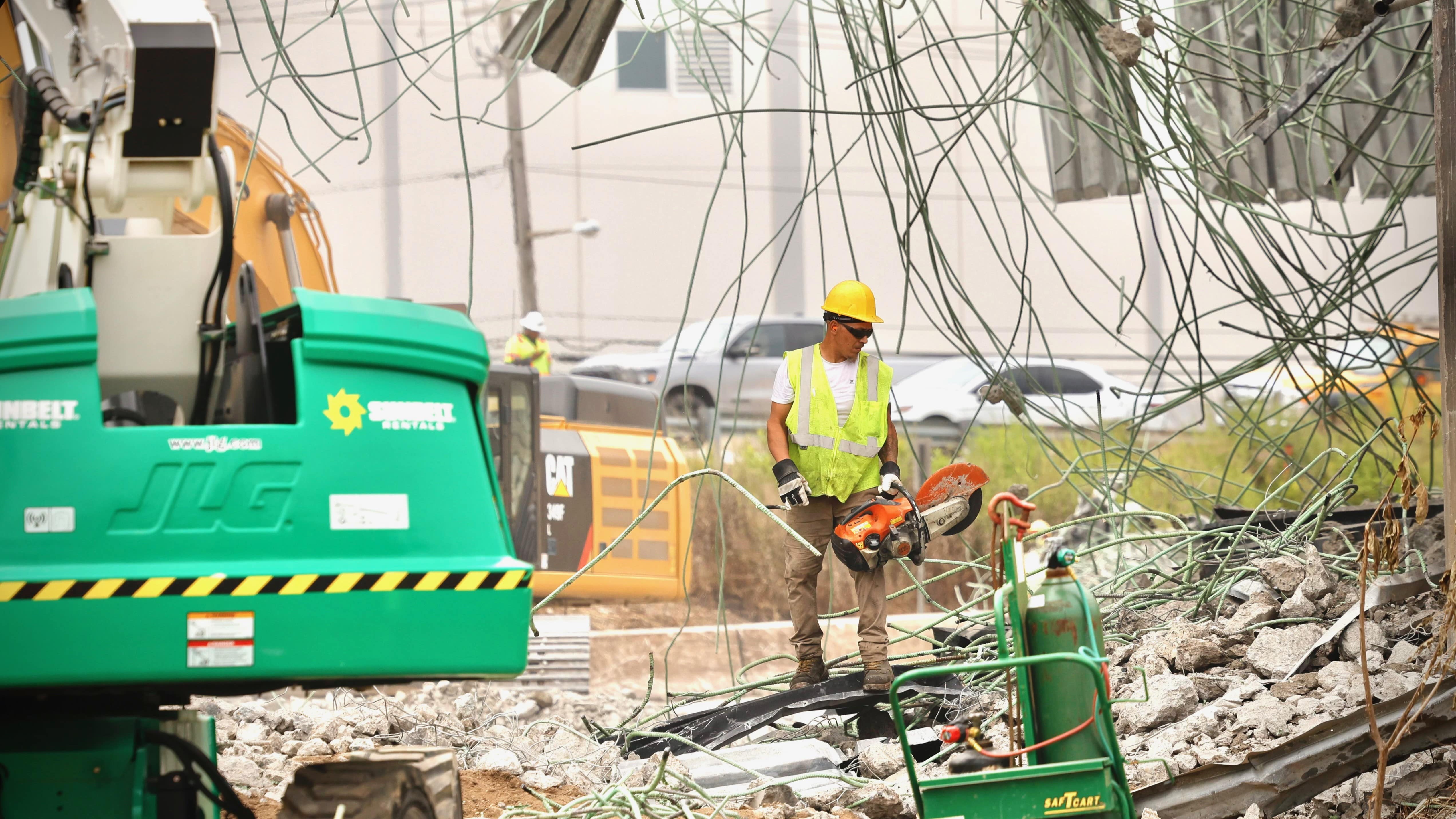 A worker wearing a yellow hard hart hat, yellow safety vest, safety gloves and black sunglasses stands amidst the rubble and uses a chainsaw to help clear debris from the site of the bridge collapse