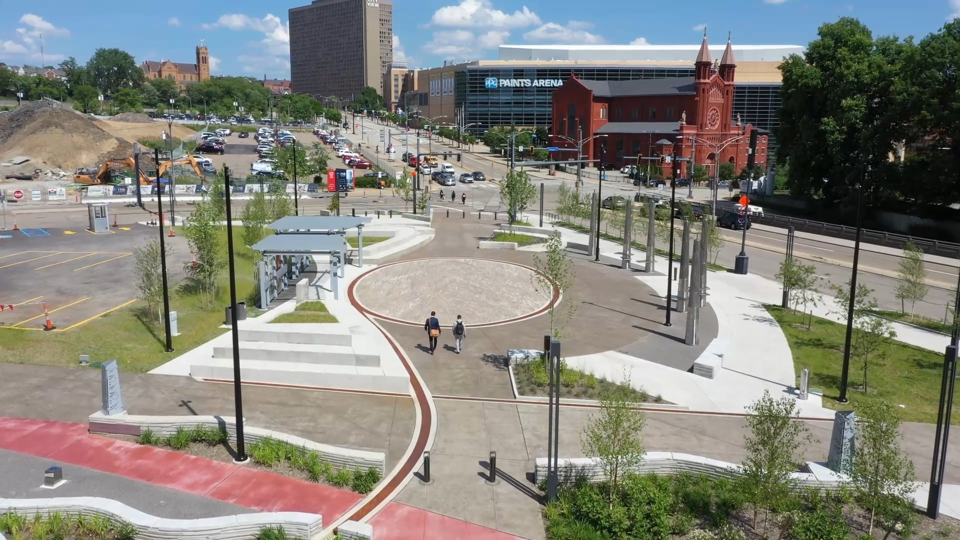 A digital rendering showing two pedestrians walking through an open space in Pittsburgh with a parking lot to the left, a roadway with vehicles on it to the right and a red brick church and the PPG Paints Arena and high-rise building in the background