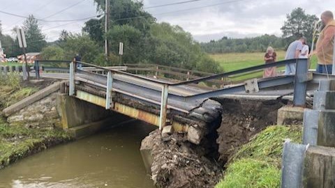 An image of a damaged and now impassable bridge with individuals standing off to the right of the bridge looking at the damage