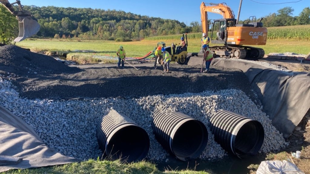 An image of a construction crew wearing hard hats and yellow safety vests building new temporary bridge with three large pipes