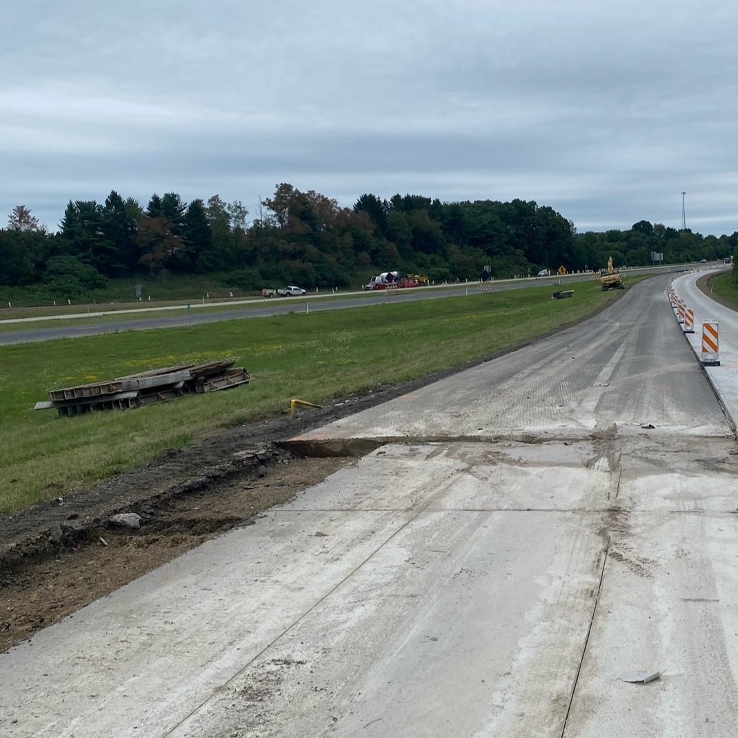 An image of a tree-lined roadway with a line of orange and white work zone barricades separating the completed right side of the roadway and the left side of the roadway being prepared for the concrete overlay